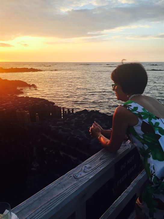 A woman leans on a wooden railing, watching the sunset over the ocean, surrounded by serene beauty.