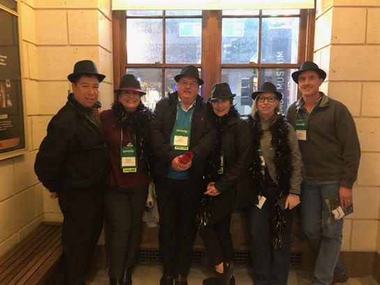 Six people dressed in hats stand smiling inside a historical building during an event.
