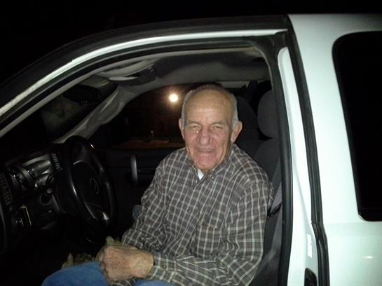 An older man smiles in his white truck at night, bathed in a soft glow.