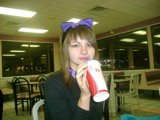 A young woman with a purple bow is enjoying a drink in a fast-food restaurant at night.