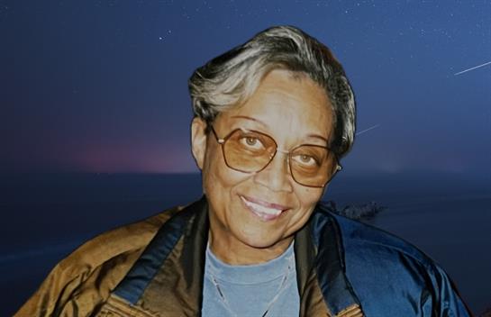 Woman smiles warmly, wearing glasses and a jacket, with a stunning ocean sunset behind her.