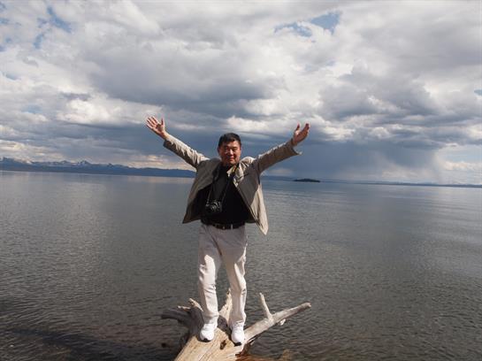A person stands on a log by the lake, arms raised, embracing the beautiful yet cloudy day.