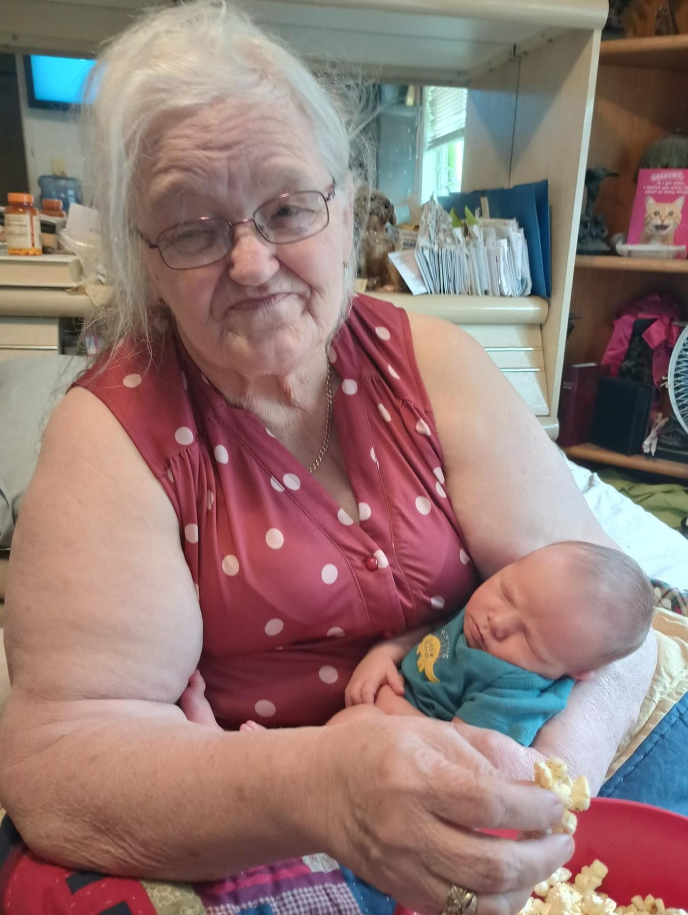 Grandmother relaxes while cradling a sleeping baby and sharing some popcorn in her home.
