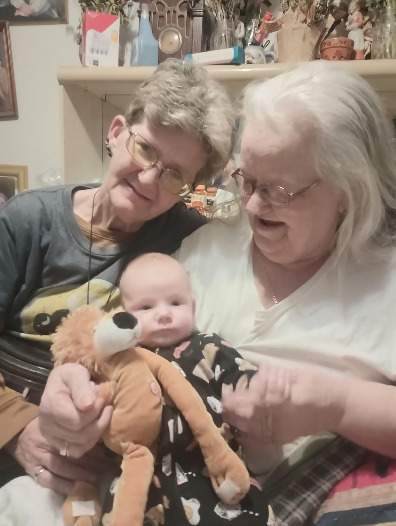 Two older women smile as they hold a baby and a stuffed animal in their laps at home.