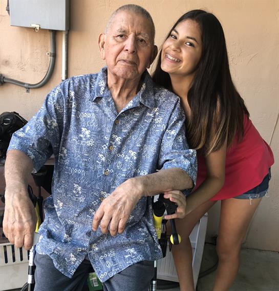 An elderly man sits in a wheelchair while a young woman stands beside him, smiling happily.