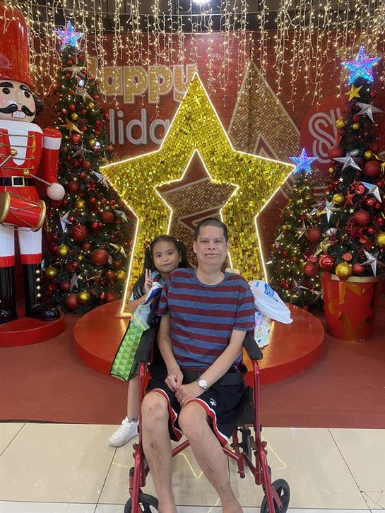 A man sits in a wheelchair with a child beside him in front of vibrant holiday decorations.