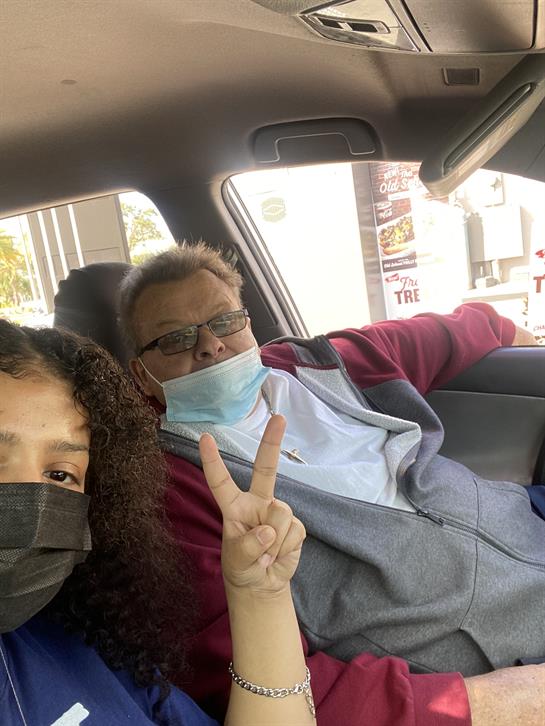 Two friends sit in a car with masks on, smiling and giving peace signs during their drive.