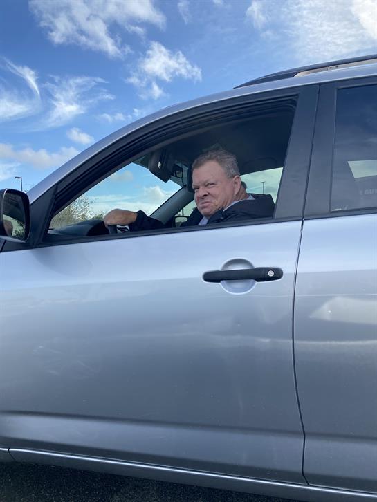 A man appears focused while driving his silver vehicle, with clouds visible overhead.