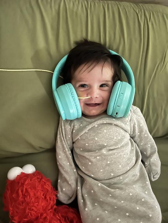 A baby smiles happily while wearing blue headphones and cuddling a red plush toy.