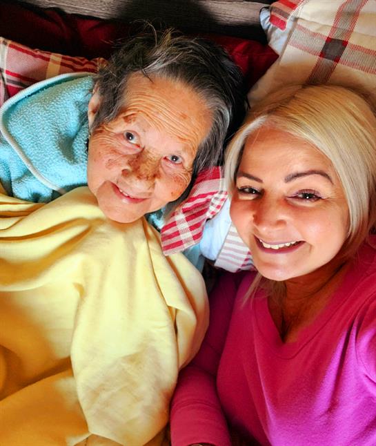 Two women are sharing a moment of joy under a cozy blanket, indoors in bright light.