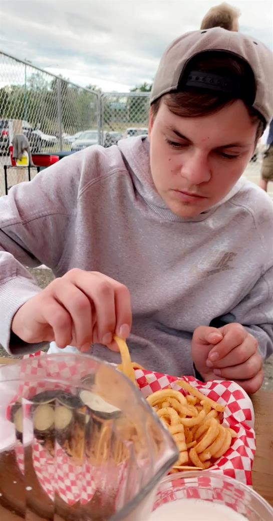A young man in a light gray hoodie focuses on his curly fries while dining outdoors.