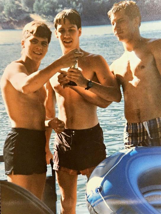 Friends gather by the lake on a warm day, holding drinks and enjoying each other's company.