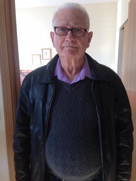 An elderly man wearing glasses and a black leather jacket poses against a plain background.