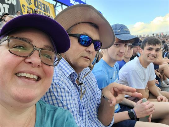 Group of four people takes a selfie while attending an outdoor event under the sun.
