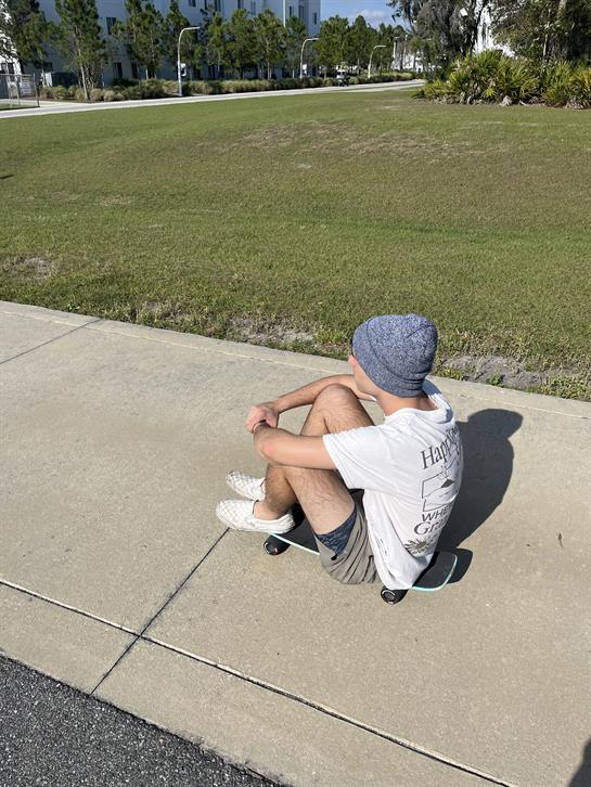 A young individual sits on a skateboard, enjoying a warm, sunny day in a park setting.