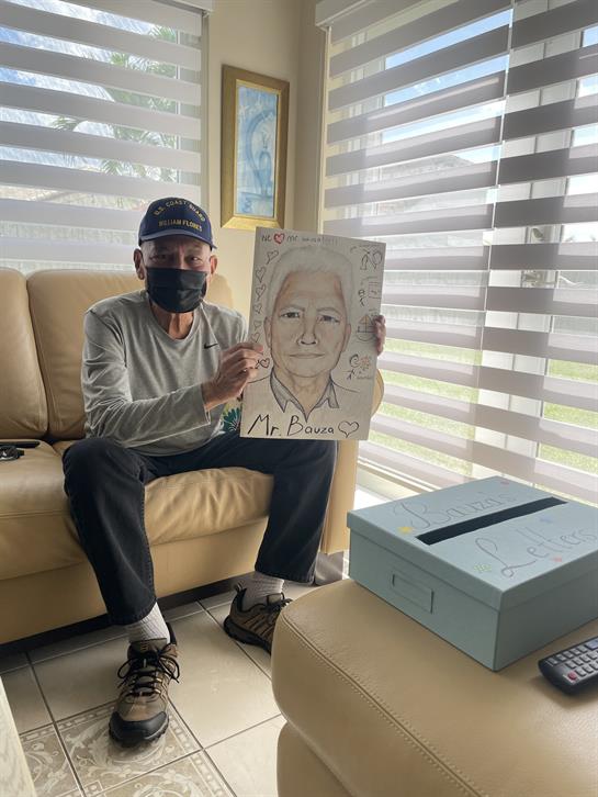 An elderly man sits on a couch, proudly displaying a drawing of a woman while wearing a face mask.