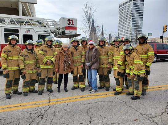 Firefighters dressed in full gear stand smiling with local residents in a city area during winter.