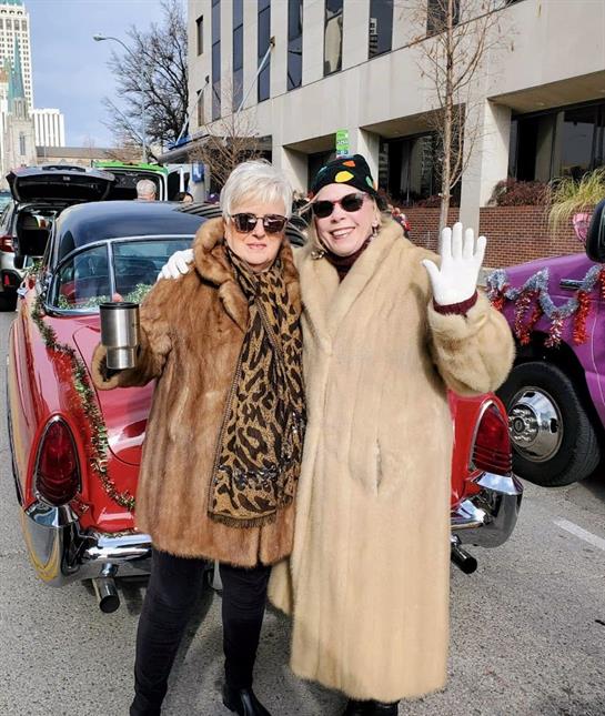 Two women pose in elegant fur coats, smiling and celebrating a winter event near vintage cars.