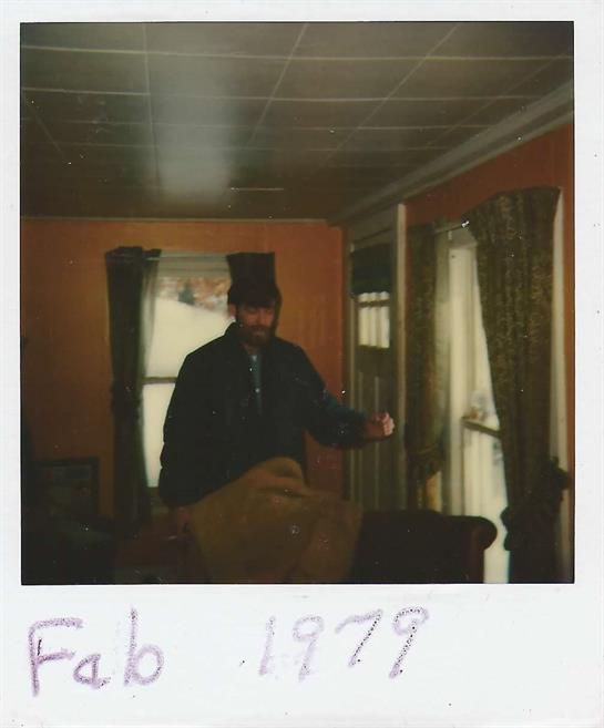 A man in a top hat stands in a cozy, vintage room, engaging with the surroundings in February 1979.