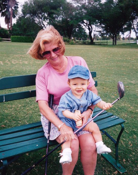 A joyful grandmother sits on a bench holding her smiling grandson, who plays with a golf club.
