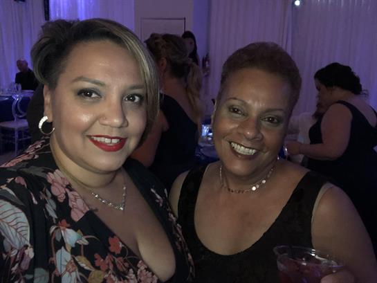 Two women are smiling and holding drinks, capturing a joyful moment at a festive gathering.