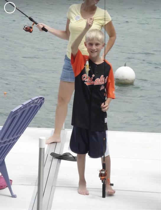 A joyful boy holds up his fishing catch as an adult poses nearby on the dock by the water.