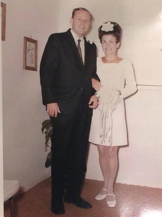 A man in a suit and woman in a white dress stand close together, smiling before an event.