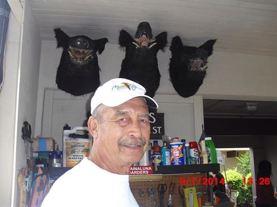An elderly man stands in a workshop, smiling in front of mounted animal heads while crafting.