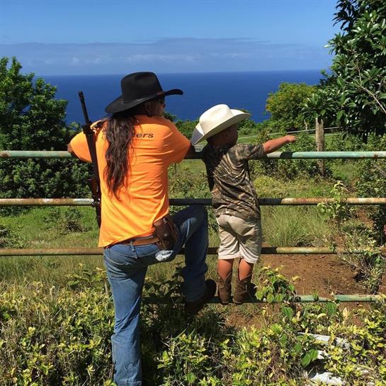 A child points toward the ocean as an adult leans against a fence, enjoying nature.