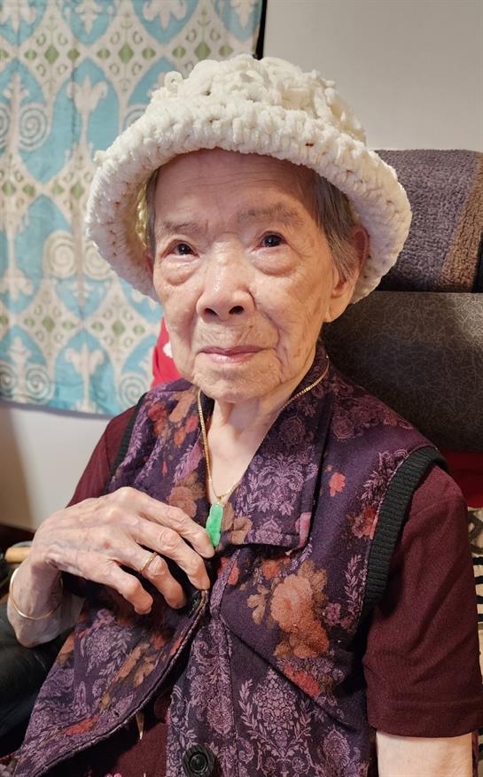 An elderly woman wearing a hat and traditional clothing sits in a chair, smiling softly.