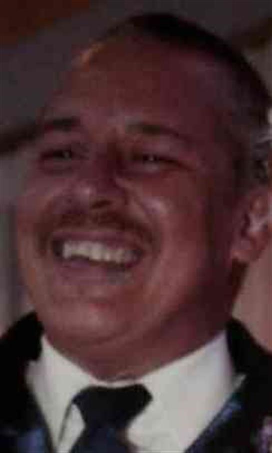 A man wearing a tuxedo smiles broadly, surrounded by joyful guests at an indoor celebration.