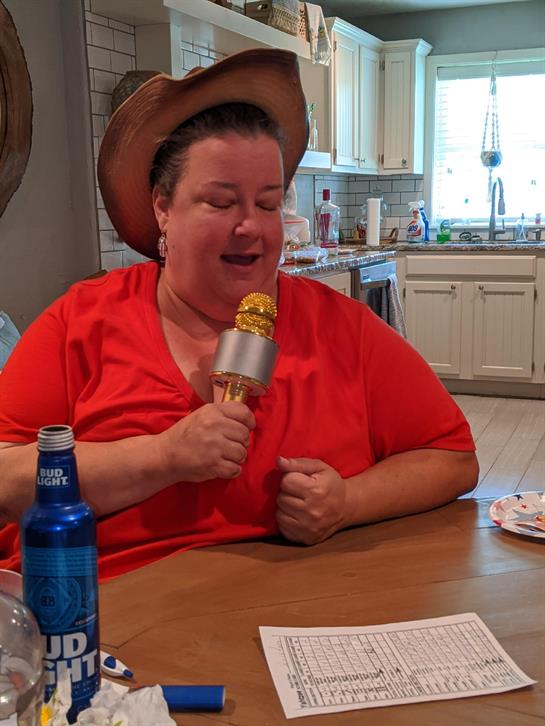 A woman in a red shirt joyfully sings into a microphone while seated at a kitchen table.