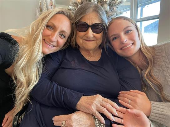 Two women share a warm embrace with an elderly woman, all smiling happily together indoors.
