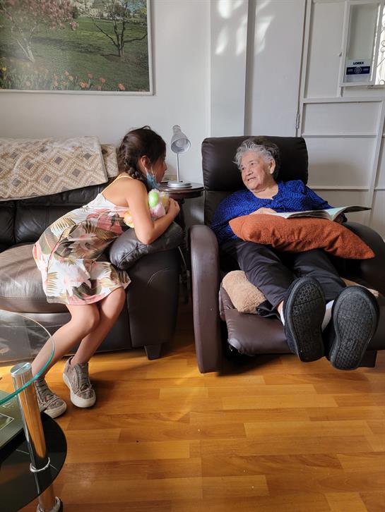 A young girl leans in to talk to an elderly man resting in a recliner chair at home.