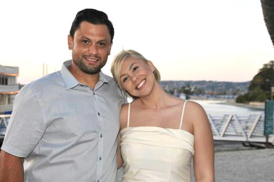 A couple poses happily by the water, smiling under the soft glow of a sunset.