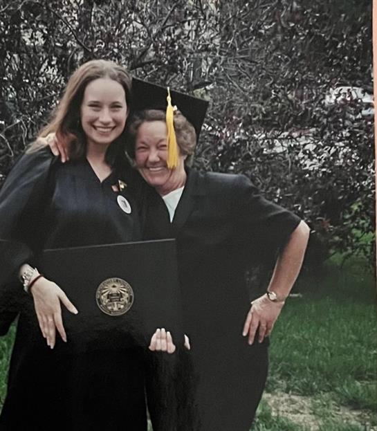 Two women celebrate graduation with smiles while holding a diploma outside in a garden.