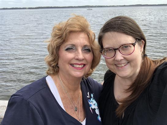 Two smiling women pose together by the waterfront, enjoying a beautiful day outdoors.