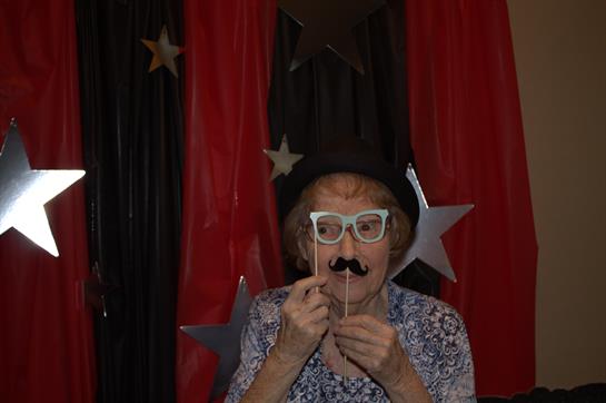 An elderly woman playfully poses with a fake mustache at a celebration.