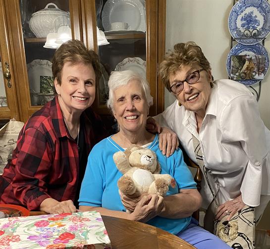 Three women smile together at a table while one holds a teddy bear, enjoying their time.