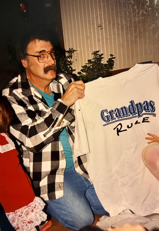 A grandfather smiles while holding a T-shirt that celebrates his role at a family event.