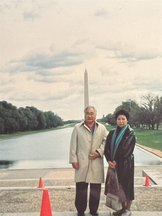 A couple stands near the reflecting pool, gazing at the Washington Monument on an overcast day.