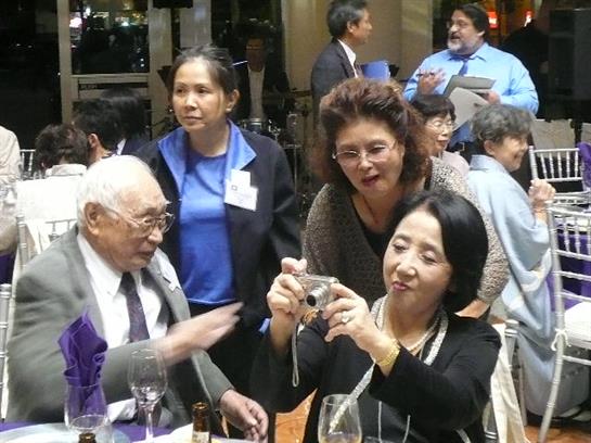 Elderly guests interact and capture moments during a lively social event in a banquet venue.