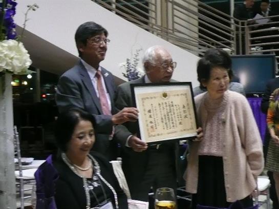 Guests at an award ceremony gather around to celebrate and display a certificate of recognition.