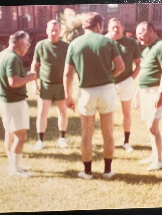 A group of six men wearing green athletic shirts gather in conversation on a sunny day.