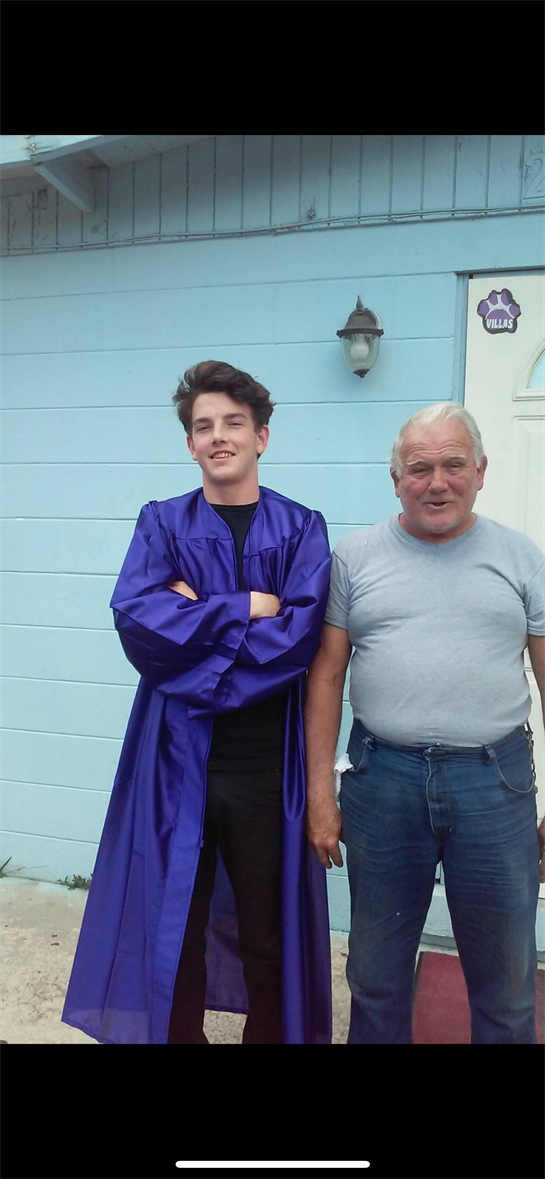 Young man in a graduation gown stands proudly beside an older gentleman.