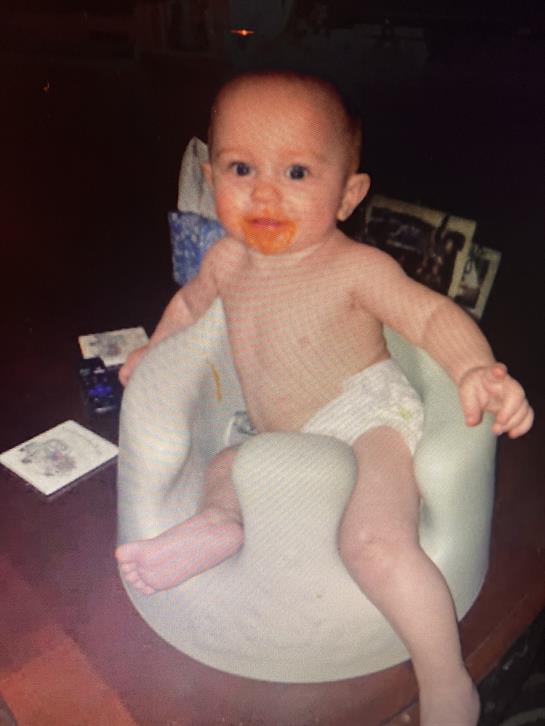 A baby in a diaper sits in a booster seat, colorful food on their mouth, at home in the afternoon.