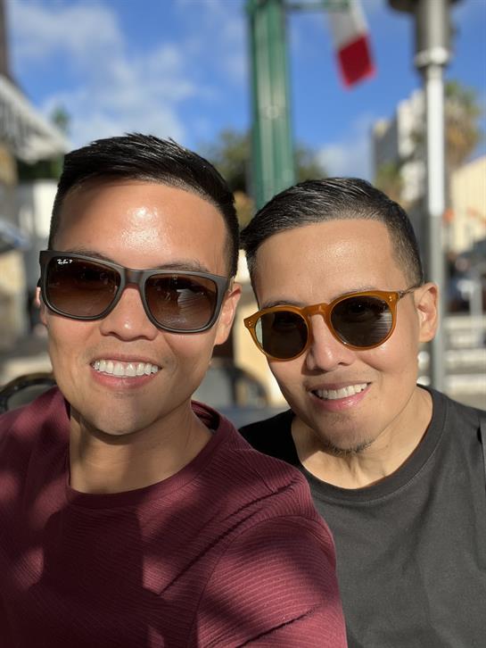 Two individuals relax outdoors on a sunny day, both wearing sunglasses and smiling joyfully.