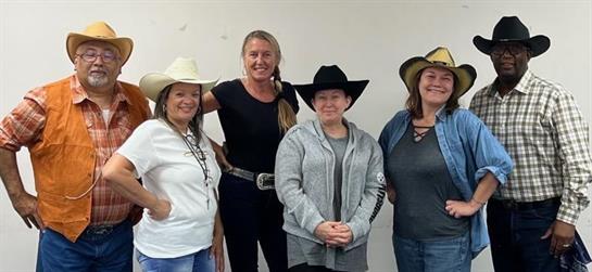 Six individuals in cowboy hats pose together, showcasing their western attire with smiles.