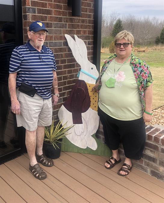 Two seniors stand next to a colorful bunny decoration outside their residence, smiling happily.
