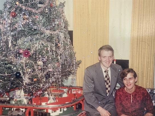 This couple poses in front of a beautifully decorated Christmas tree with a train set.